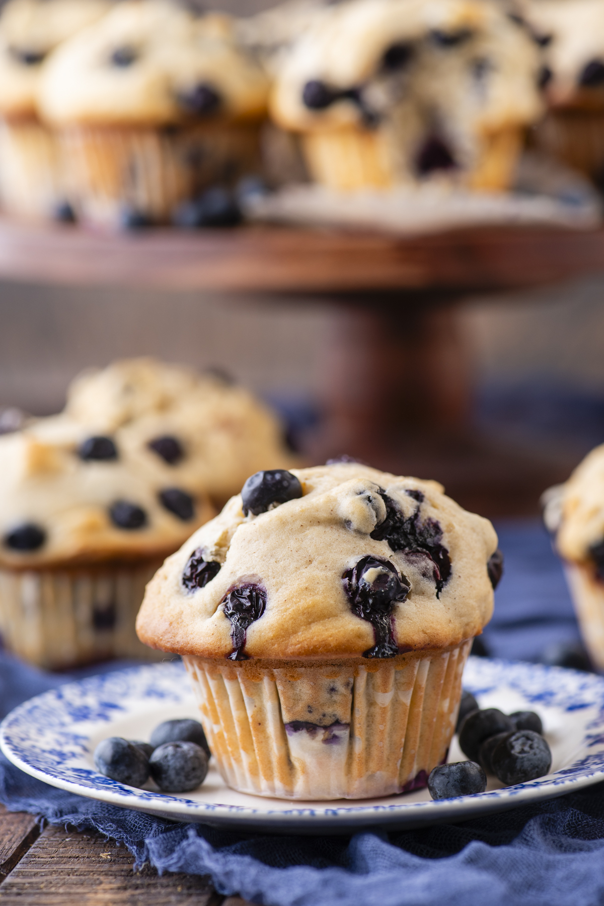 A freshly baked blueberry muffin sits on a blue and white plate with whole blueberries, with more muffins on a cake stand in the background. The muffin is golden brown and studded with juicy blueberries.