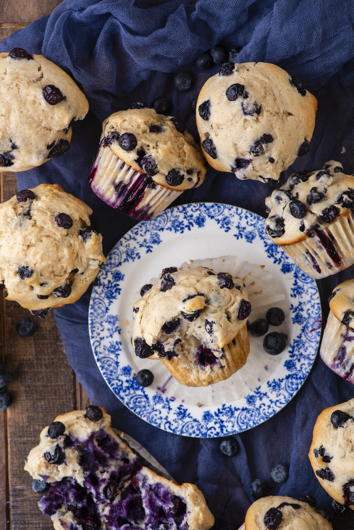 Overhead view of several blueberry muffins, some on a blue-patterned plate and others on a wooden surface and blue cloth, with loose blueberries scattered around. One muffin is cut open, showing the blueberry-filled center.
