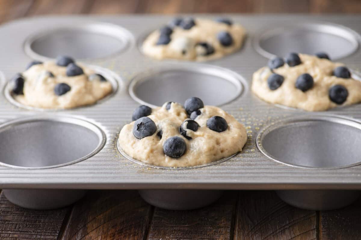 A metal muffin tin with several cups filled with blueberry muffin batter, some topped with fresh blueberries, resting on a wooden surface. A few cups are empty.