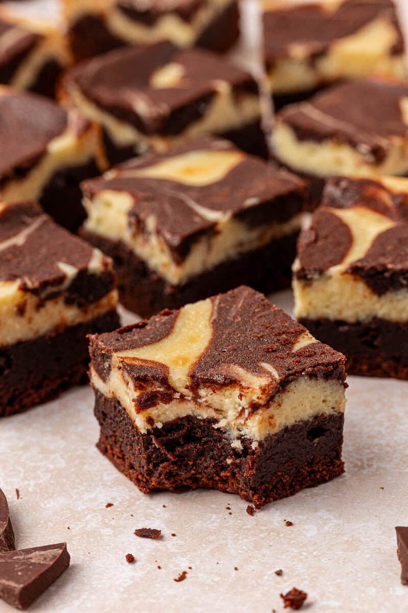 A close-up of marbled chocolate cheesecake brownies, with one brownie in the center showing a bite taken out. The brownies have a swirled chocolate and cream cheese pattern on top.