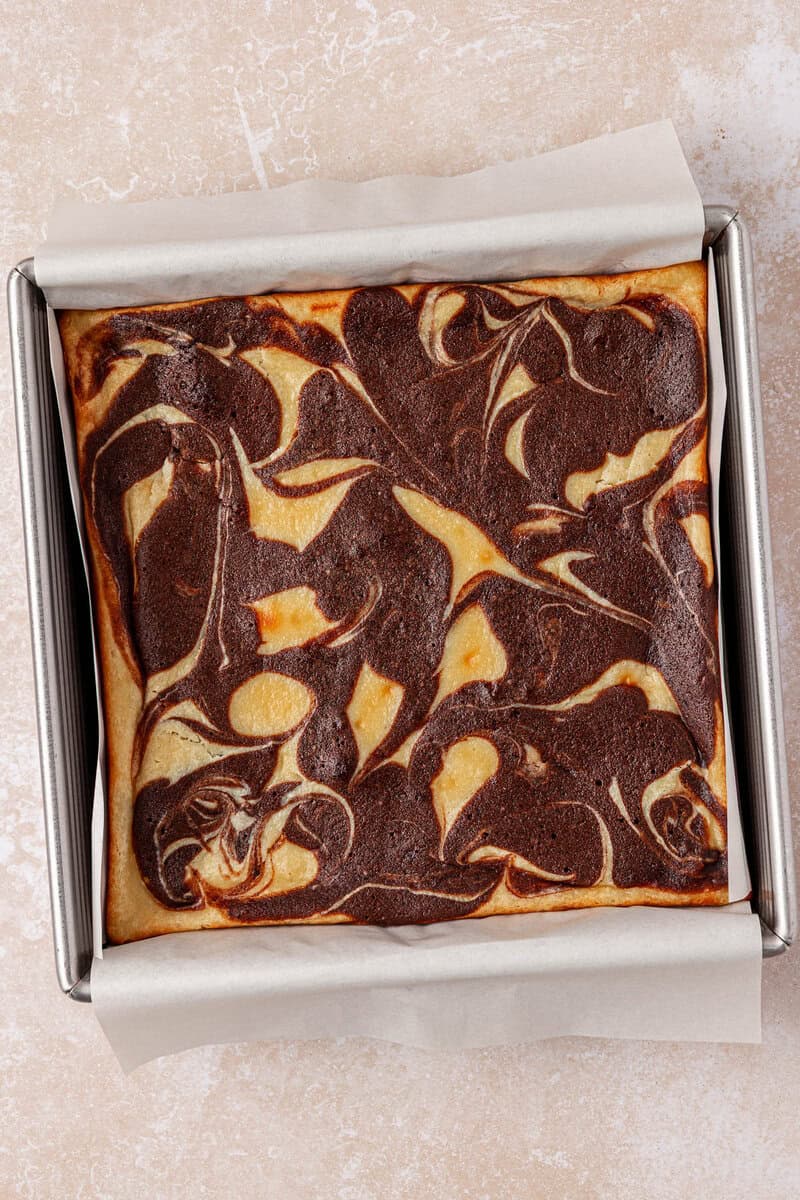 A square pan of freshly baked cheesecake brownies with a marbled chocolate and cream cheese swirl pattern on top, sitting on parchment paper.
