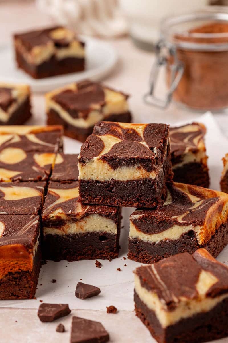 A close-up of several stacked chocolate brownies with a creamy cheesecake swirl, arranged on parchment paper with chocolate pieces nearby. Some brownies are in the background on a white plate.