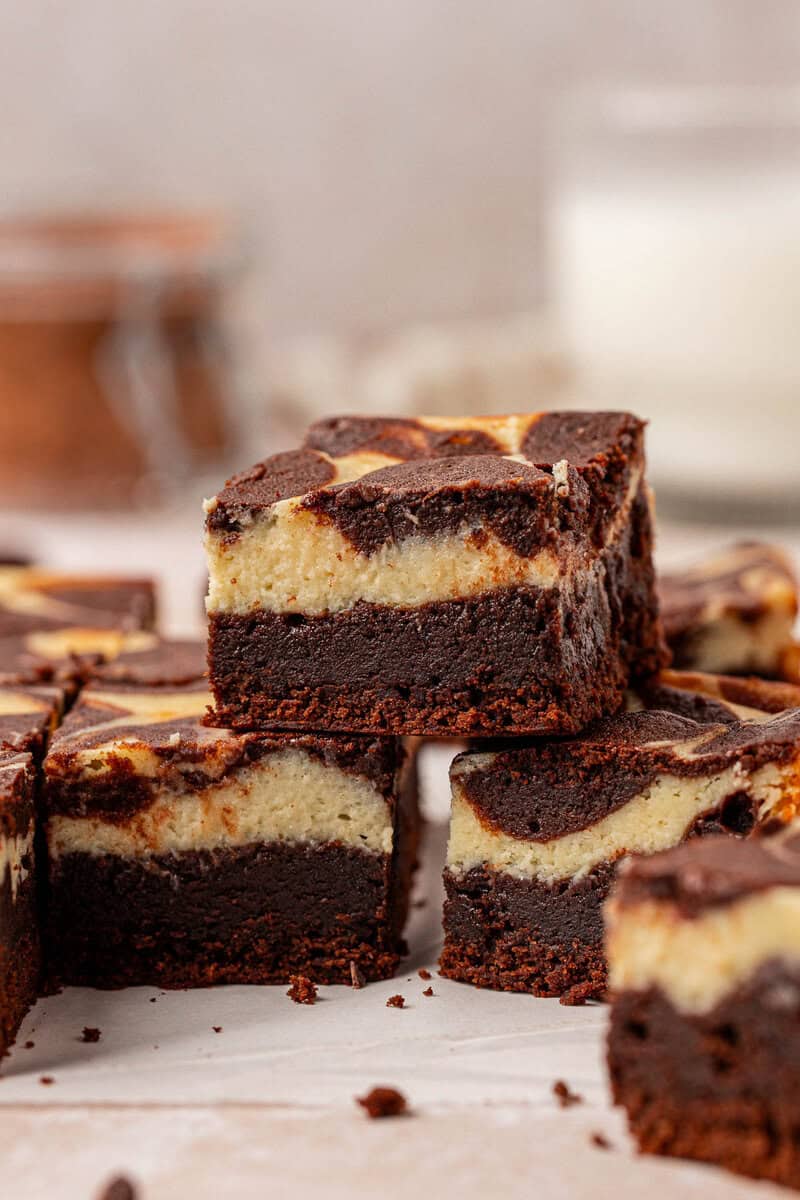 Close-up of stacked chocolate brownies with a creamy cheesecake swirl on top, arranged on a light surface. The texture of both the brownie and cheesecake layers is clearly visible.