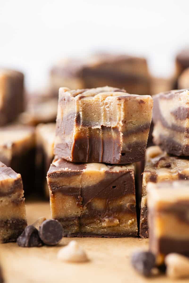 Close-up of stacked squares of chocolate and peanut butter fudge with visible swirled layers, set on a wooden surface with scattered chocolate and peanut butter chips in the foreground.