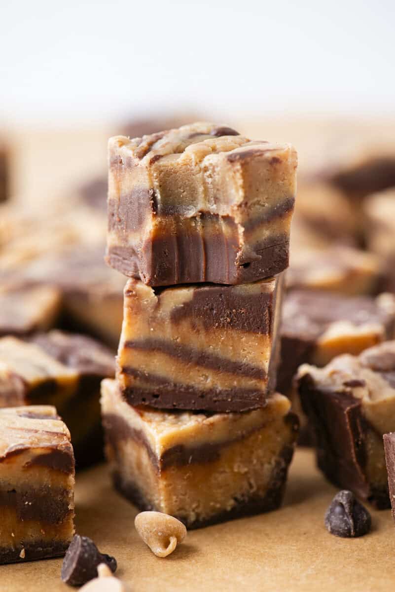 Three pieces of marbled chocolate and peanut butter fudge are stacked on top of each other, with more fudge pieces and chocolate chips scattered in the background on a brown surface.
