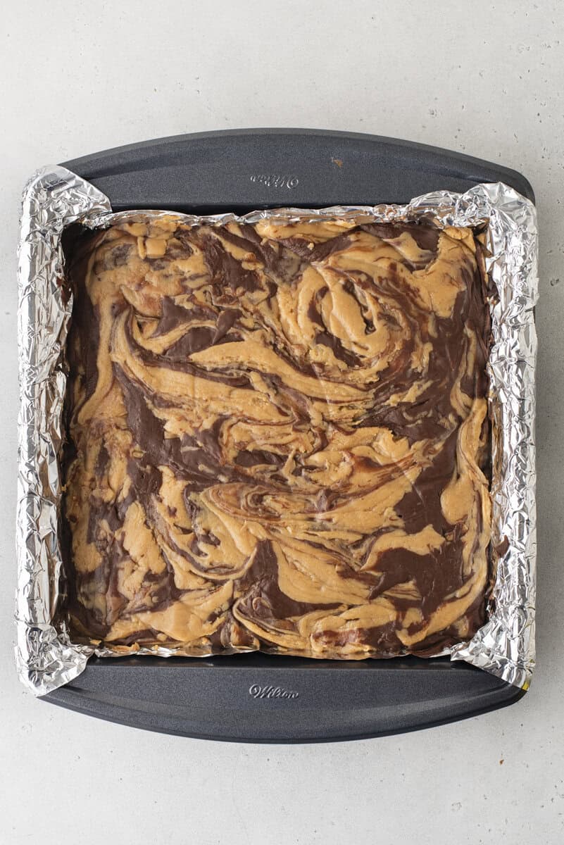 A square baking pan lined with foil holds a batch of brownies with swirled chocolate and peanut butter patterns on top, viewed from above.