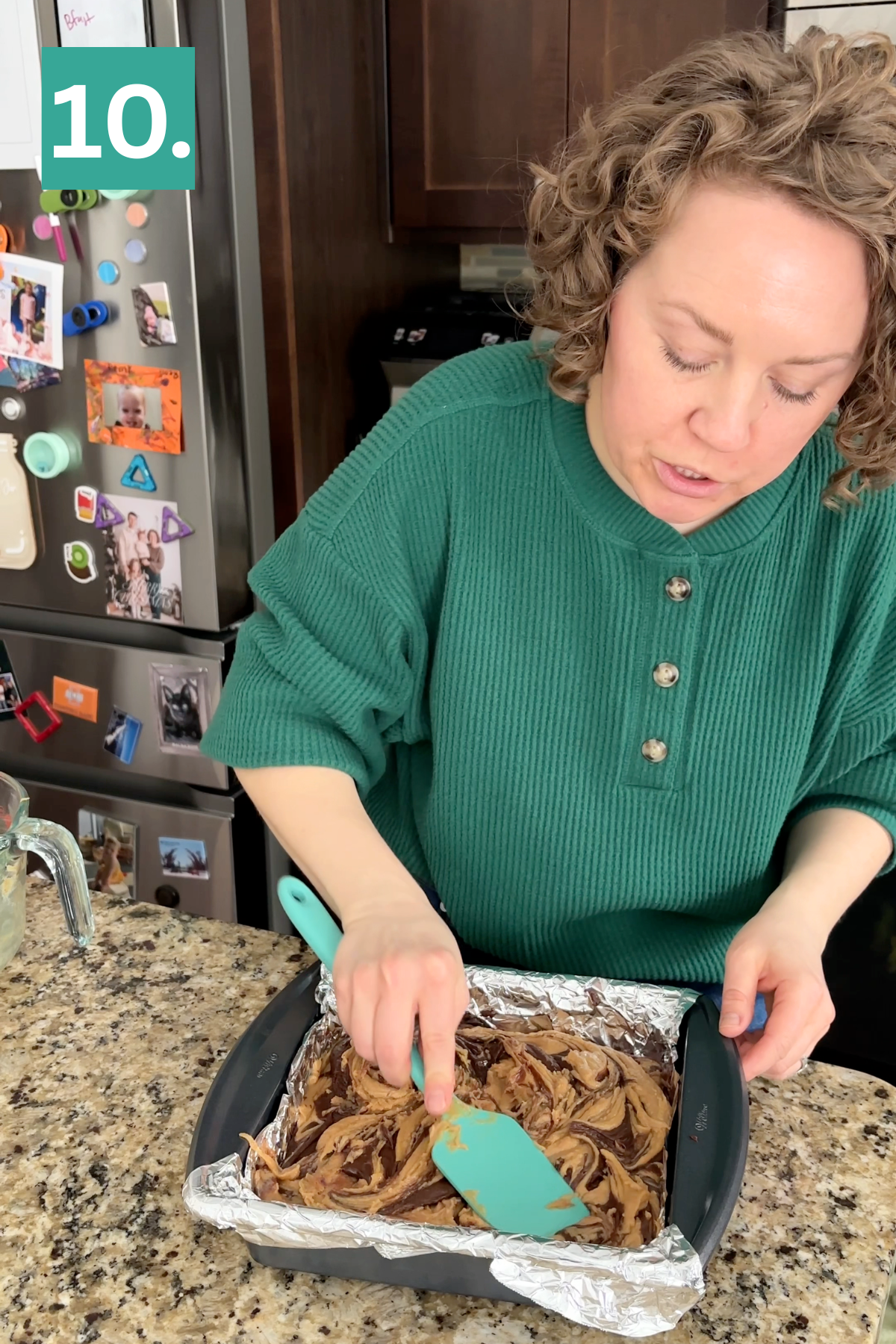 A woman in a green shirt spreads marbled batter in a foil-lined square baking pan with a spatula on a kitchen counter. A fridge with magnets and photos is in the background. A green box with 10. is in the top left corner.
