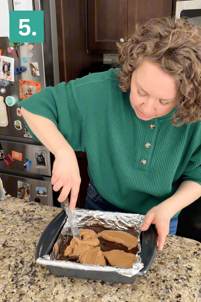 A person in a green sweater spreads chocolate frosting on a dessert in a foil-lined baking pan on a granite countertop. A fridge with photos and magnets is in the background. A green box with the number 5 is in the top left corner.