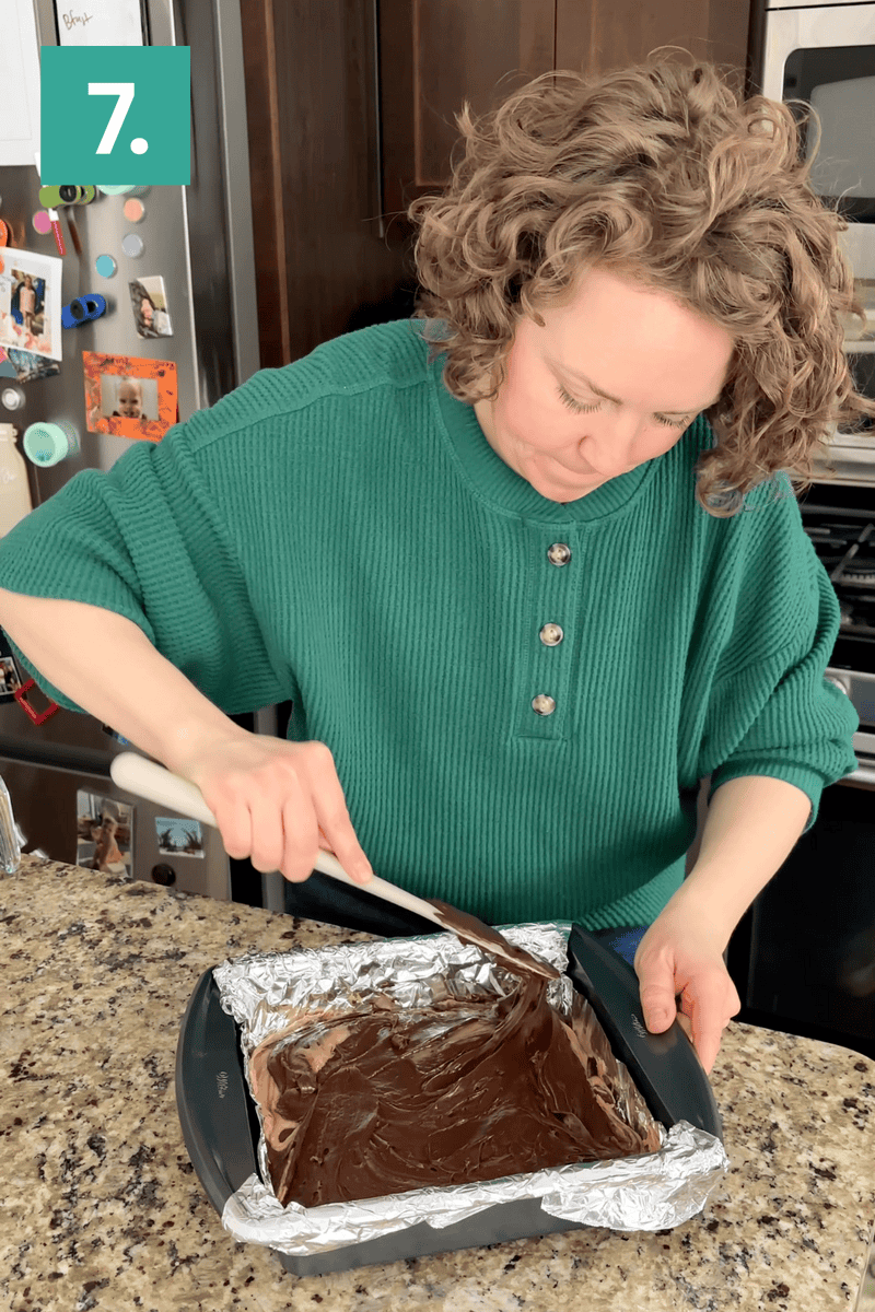 A woman in a green sweater spreads chocolate over a foil-lined pan on a granite kitchen counter. Step 7 is indicated in the top left corner. Refrigerator and cabinets are visible in the background.