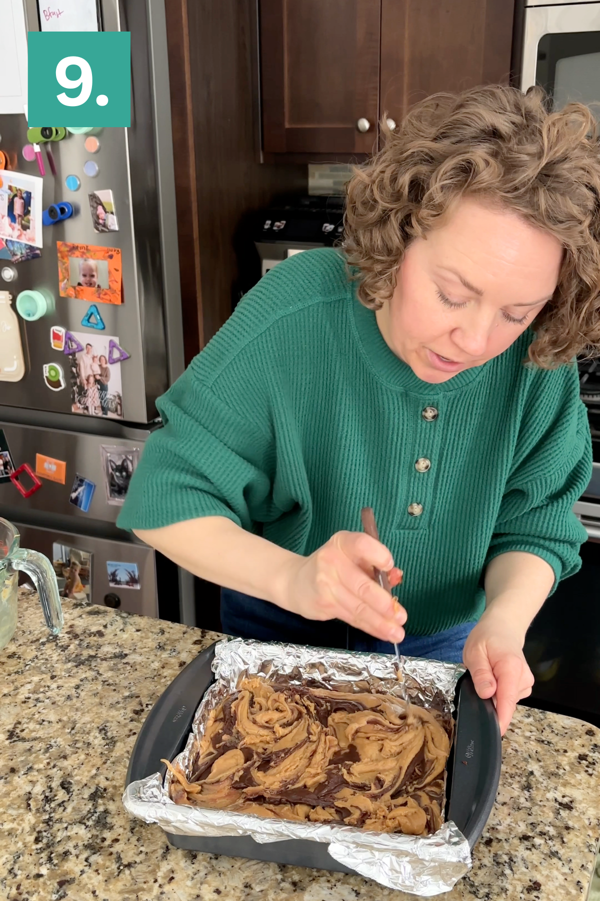 A woman in a green sweater swirls batter in a foil-lined baking pan on a kitchen counter. A refrigerator with photos and magnets is in the background, and a number 9 appears in the top left corner.