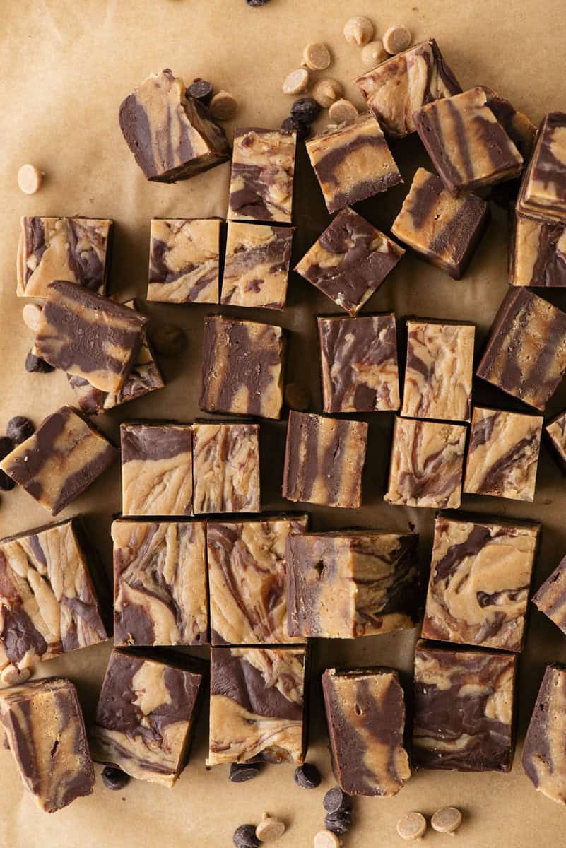Squares of marbled chocolate and peanut butter fudge are arranged on brown parchment paper, with a few scattered chocolate and peanut butter chips around them.