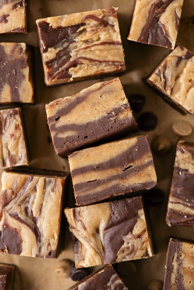 Squares of marbled chocolate and peanut butter fudge are arranged on a brown surface. The fudge has swirled layers of dark brown chocolate and light brown peanut butter. A few chocolate and peanut butter chips are scattered nearby.