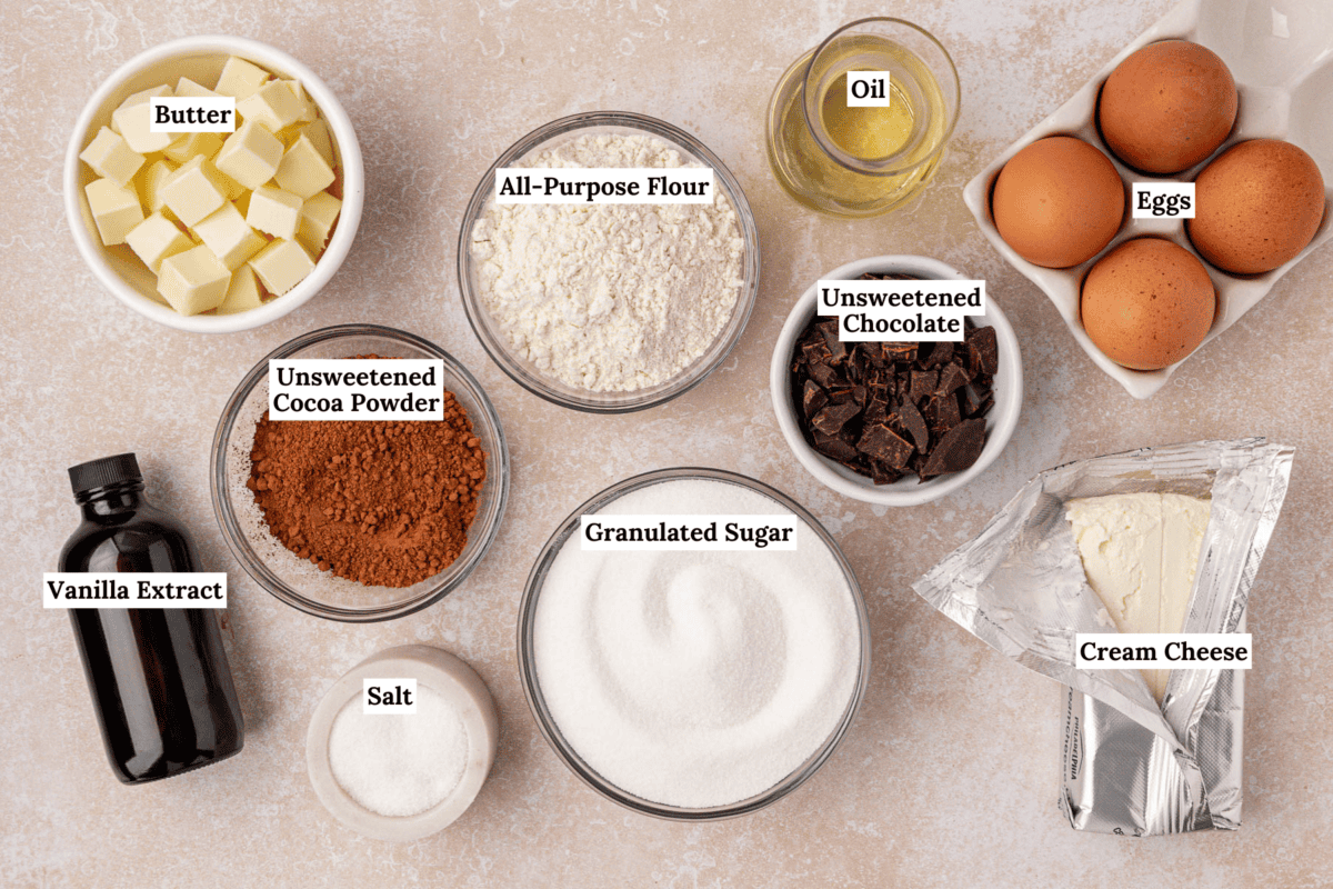 A top-down view of labeled baking ingredients in bowls and containers, including butter, flour, eggs, oil, unsweetened chocolate, cocoa powder, cream cheese, granulated sugar, vanilla extract, and salt.