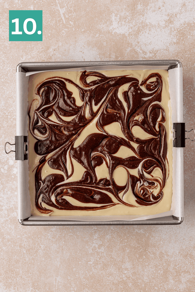 A square baking pan filled with swirled chocolate and vanilla batter sits on a light surface. The pan is lined with parchment paper, and a green box with the number 10 is in the top left corner.