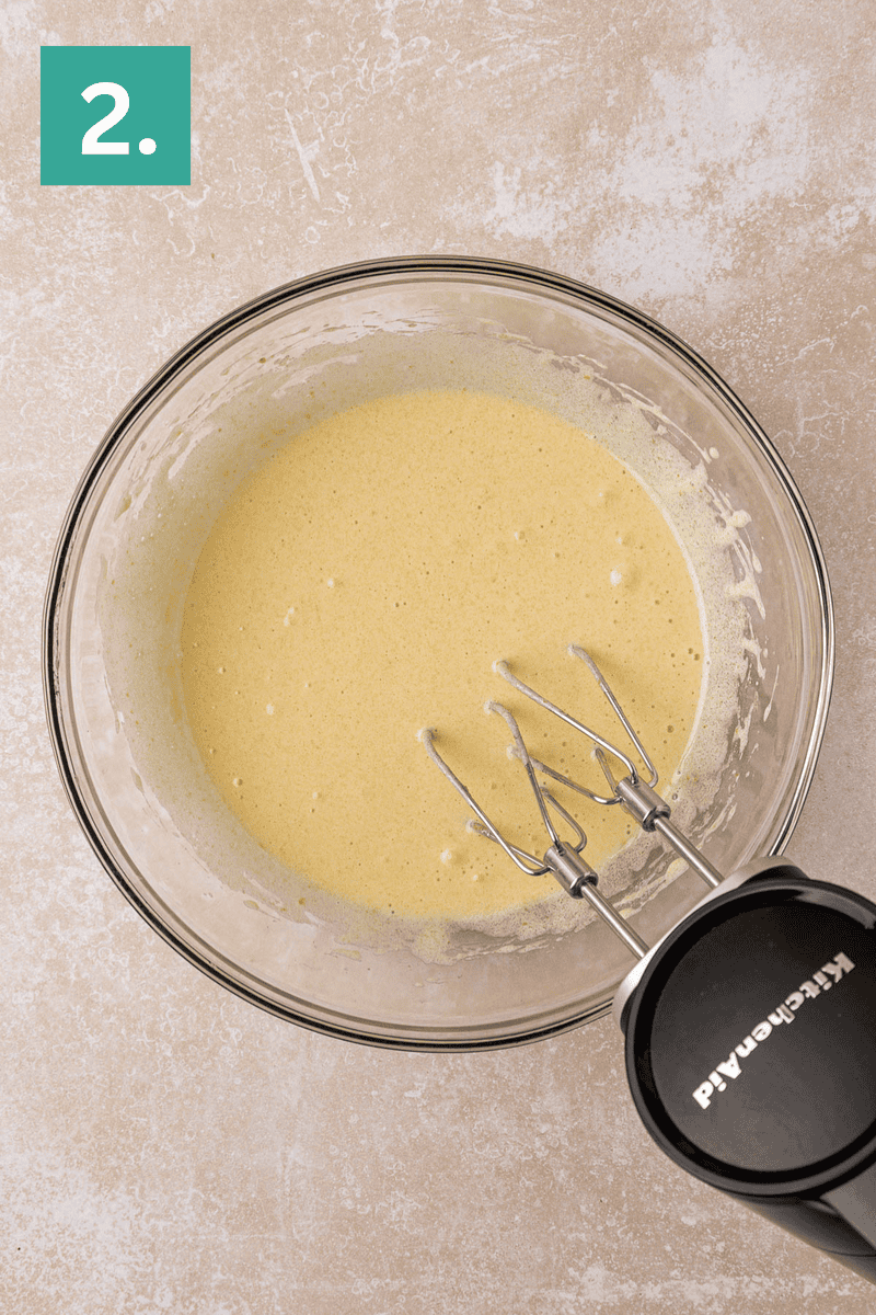 A glass mixing bowl filled with a light yellow batter is shown on a beige surface. An electric hand mixer with beaters is resting in the bowl. A green square with the number 2. is in the top left corner.