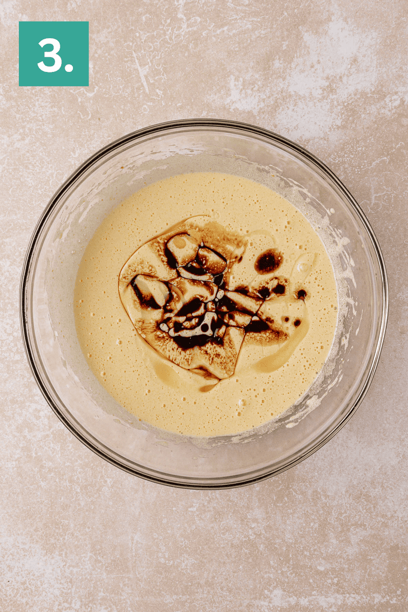 A glass bowl filled with a yellow batter and swirls of vanilla extract on top, placed on a light textured surface. A green box with the number 3 is in the top left corner.