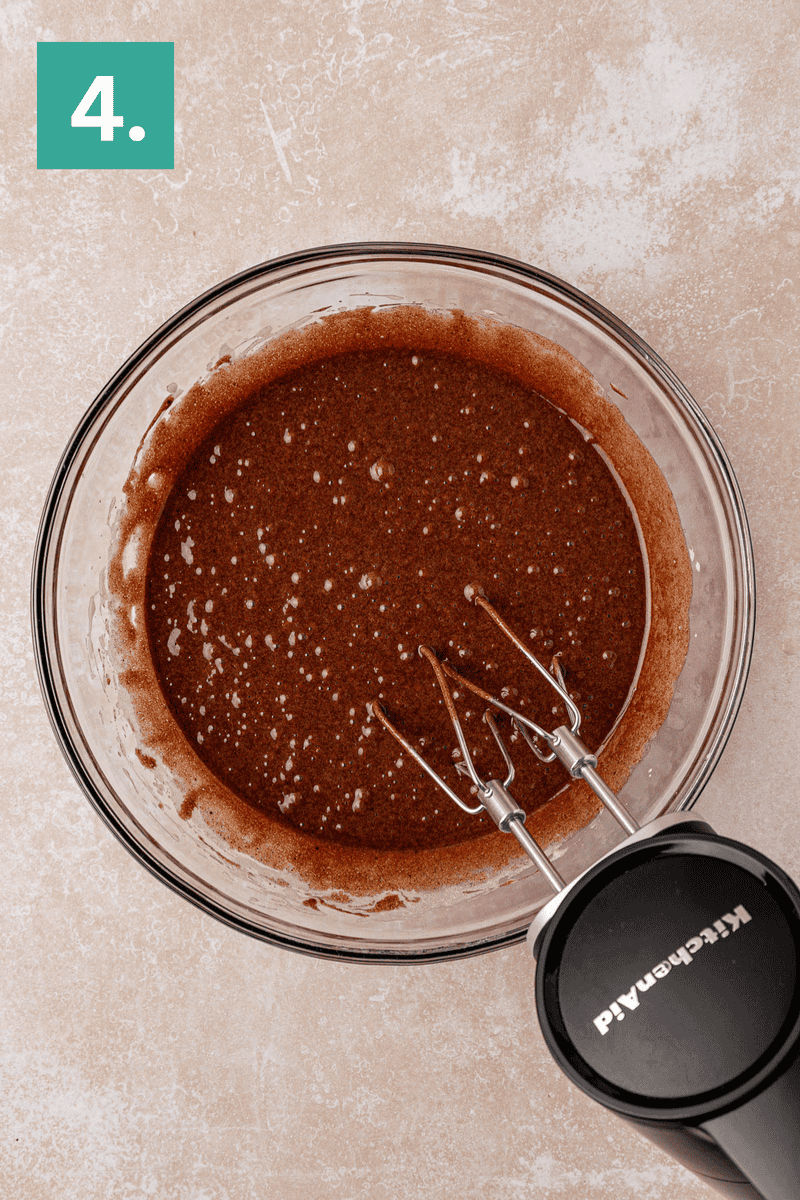 A glass bowl filled with chocolate batter sits on a light countertop. An electric hand mixer with two beaters rests in the bowl. A green box with the number 4. is in the top left corner.