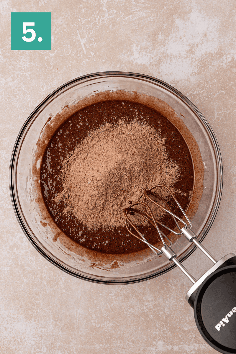A glass bowl filled with brownie batter and dry cocoa powder sits on a light surface. An electric hand mixer is placed in the bowl, ready to mix. The image is labeled with a green box containing the number 5.