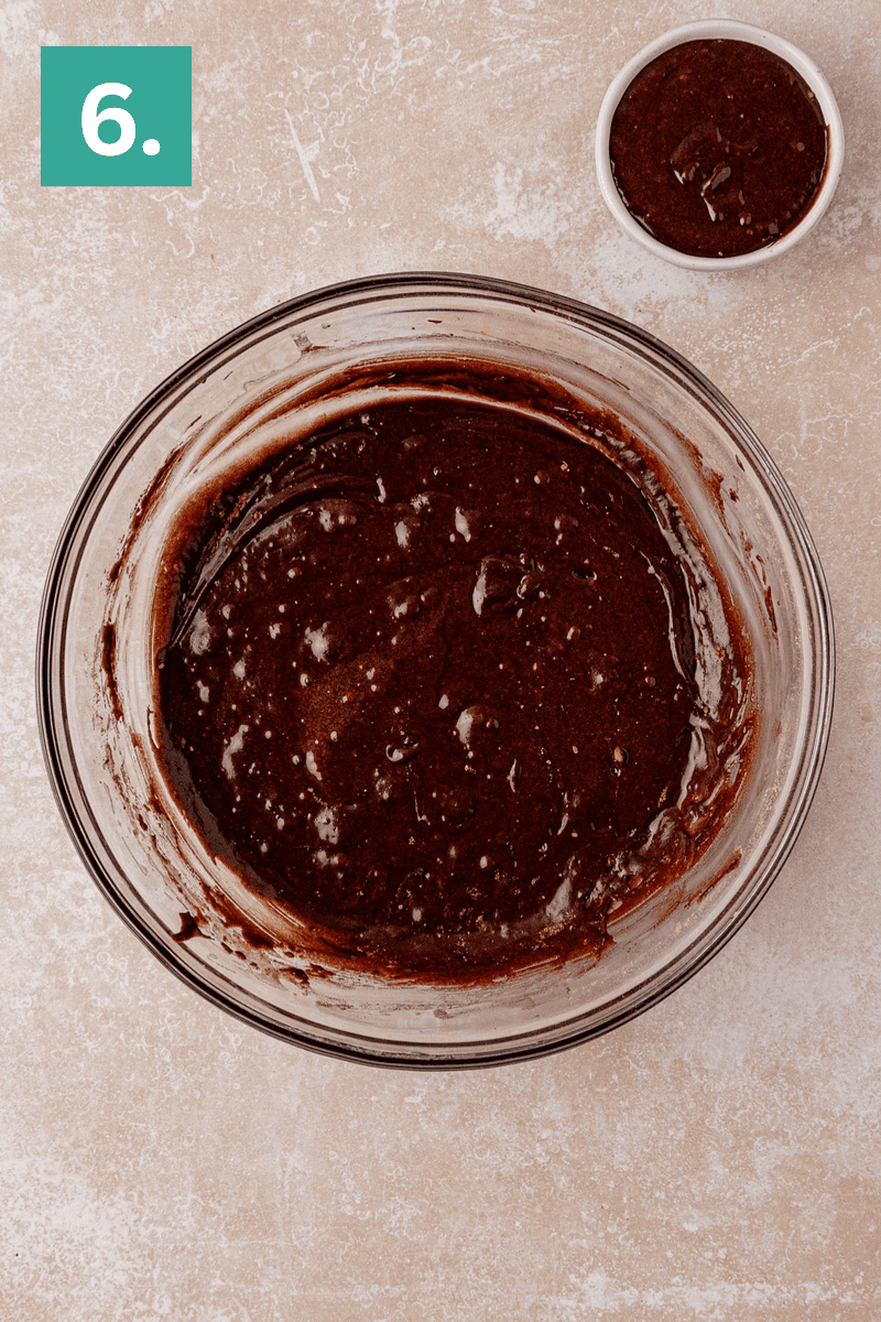 A large glass bowl filled with thick chocolate batter sits on a light countertop. A smaller bowl with more chocolate batter is in the upper right corner. A green square with the number 6 is in the upper left corner.