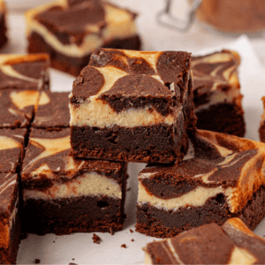Several cheesecake brownies with marbled chocolate and cream cheese tops are stacked and arranged on a white surface, showcasing their rich, fudgy layers and creamy swirls.
