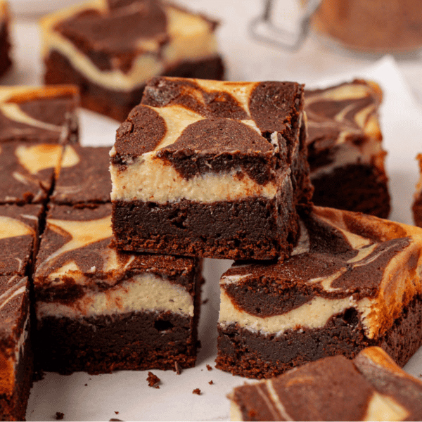 Several cheesecake brownies with marbled chocolate and cream cheese tops are stacked and arranged on a white surface, showcasing their rich, fudgy layers and creamy swirls.