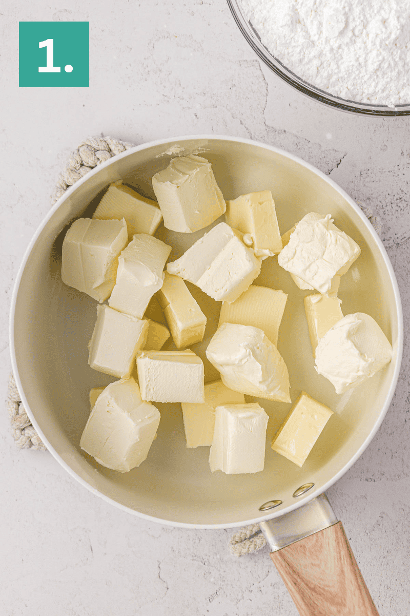A saucepan filled with cubed butter sits on a countertop next to a bowl of powdered sugar. A green box with the number 1 is in the top left corner, indicating the first step in a recipe.
