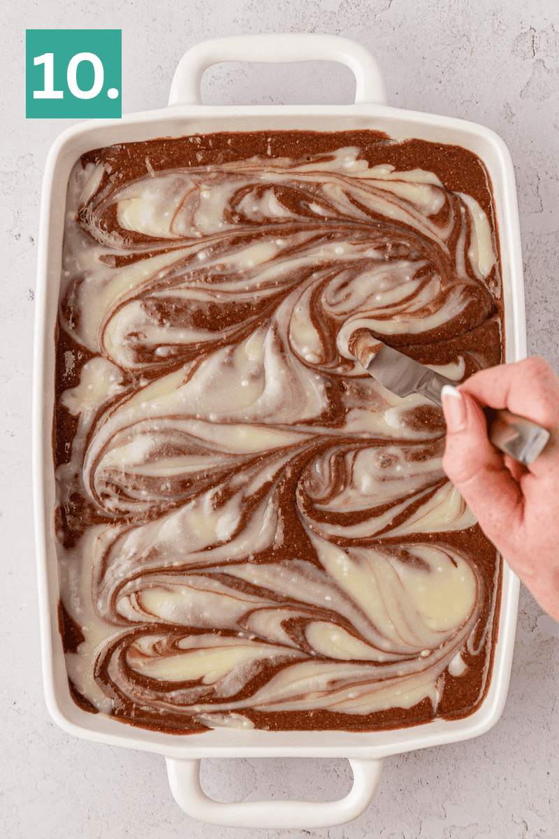 A hand holds a fork, swirling cream cheese into chocolate brownie batter in a rectangular white baking dish; a green box with the number 10 is in the top left corner.