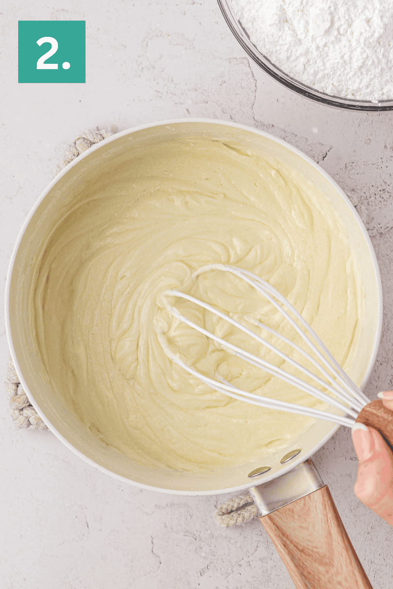 A hand holding a whisk stirs a creamy batter in a saucepan. A bowl of flour is visible in the corner. A green box with the number 2 is in the top left.