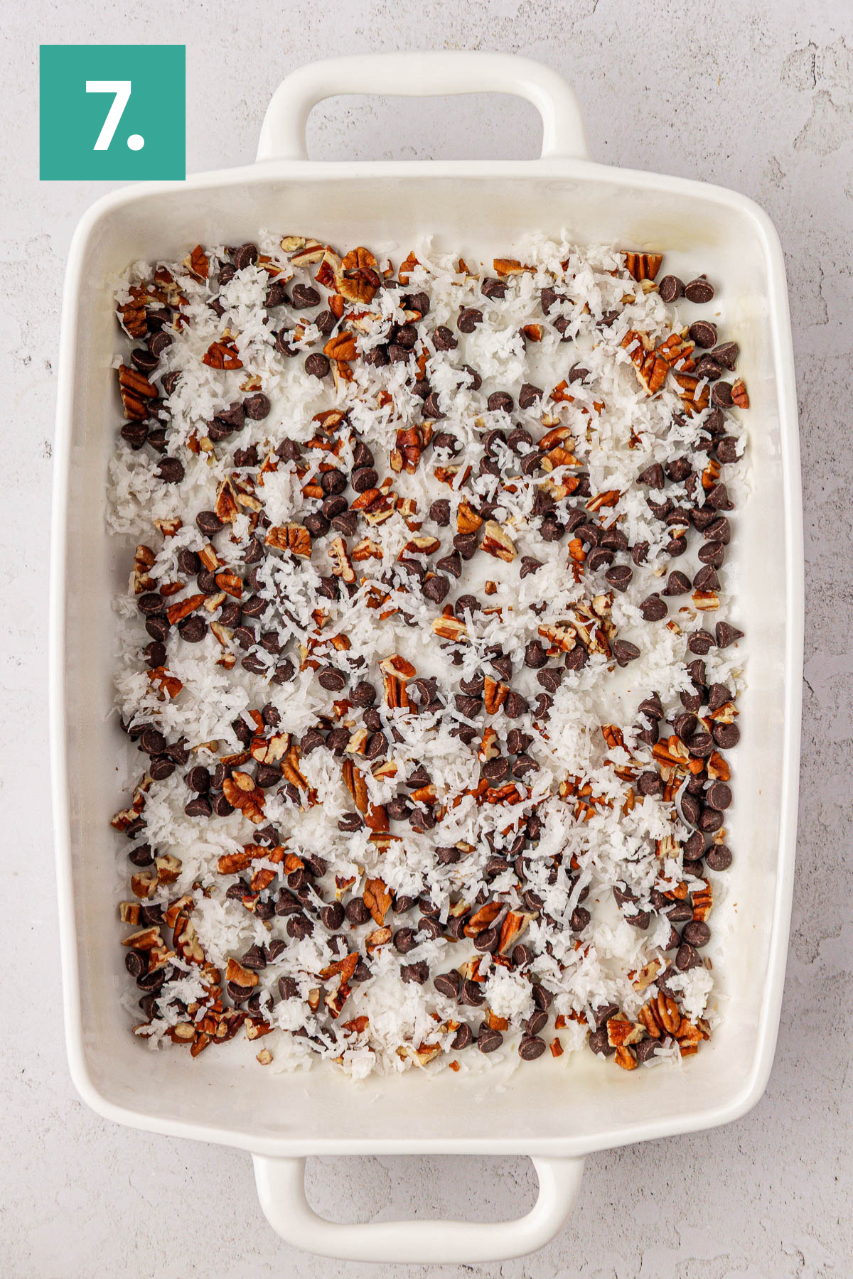 A white rectangular baking dish filled with layers of shredded coconut, chocolate chips, and chopped pecans, on a light surface. A green square in the top left corner shows the number 7.