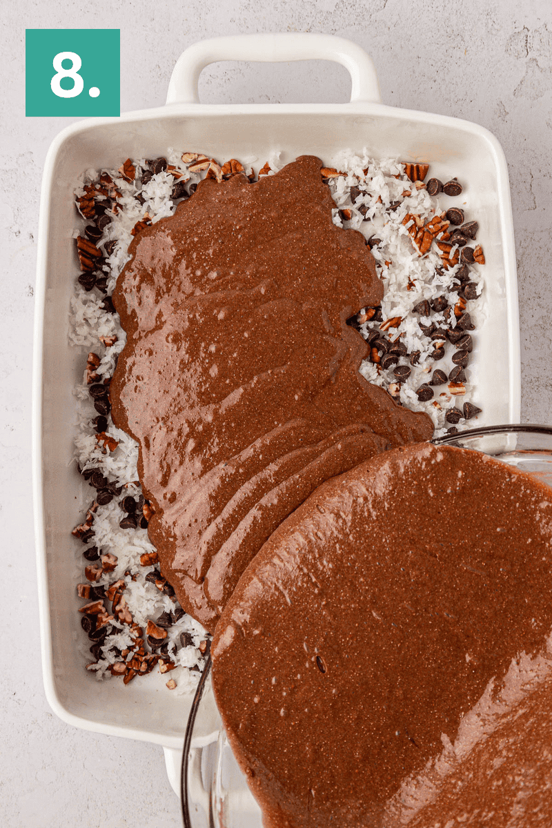 Brownie batter is being poured from a glass bowl into a white baking dish lined with shredded coconut, chocolate chips, and chopped pecans. A green box with the number 8 is in the upper left corner.