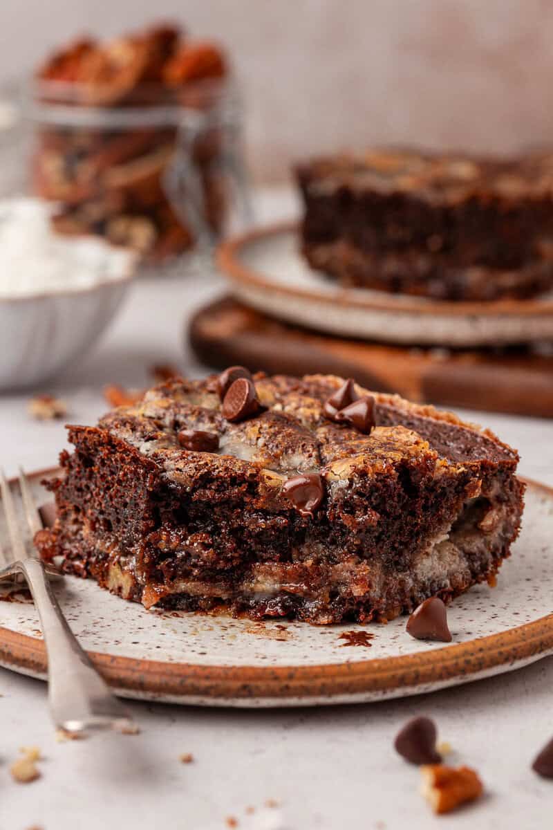 A close-up of a slice of chocolate and banana bread with chocolate chips on a ceramic plate, accompanied by a fork, with another slice and jars blurred in the background.