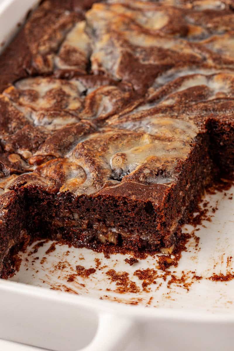 A close-up of brownies with a creamy swirled topping in a white baking dish, with a corner piece cut out, revealing the moist, chocolatey texture inside.