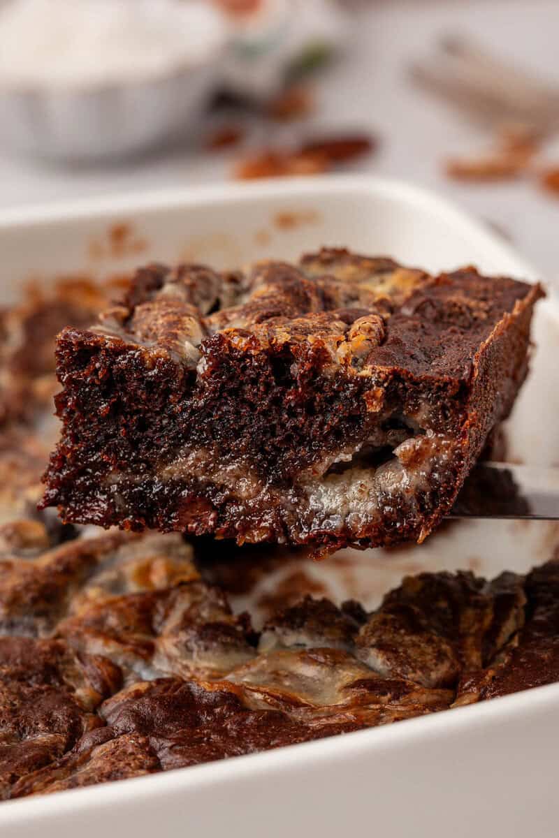 A close-up of a gooey, marbled chocolate and cream cheese brownie being lifted from a baking dish, showing a rich and fudgy texture.