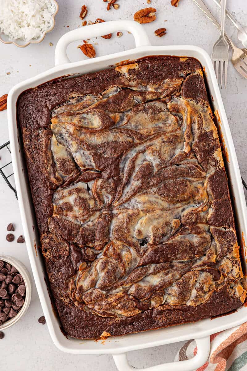 A rectangular baking dish filled with chocolate brownies swirled with a light cream cheese mixture, surrounded by a fork, pecan halves, chocolate chips, and a dish of shredded coconut.