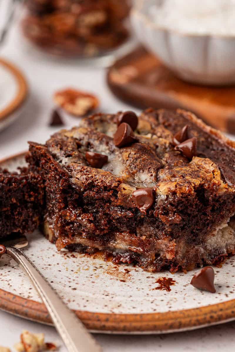 A close-up of a partially eaten gooey chocolate brownie topped with chocolate chips on a speckled ceramic plate, with a fork and scattered chocolate chips nearby.
