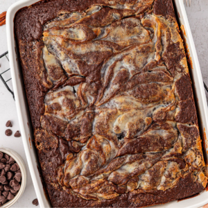 A baked chocolate brownie dessert in a rectangular white dish, featuring a swirled pattern of cream cheese on top. A bowl of chocolate chips and a wire rack are partially visible nearby.