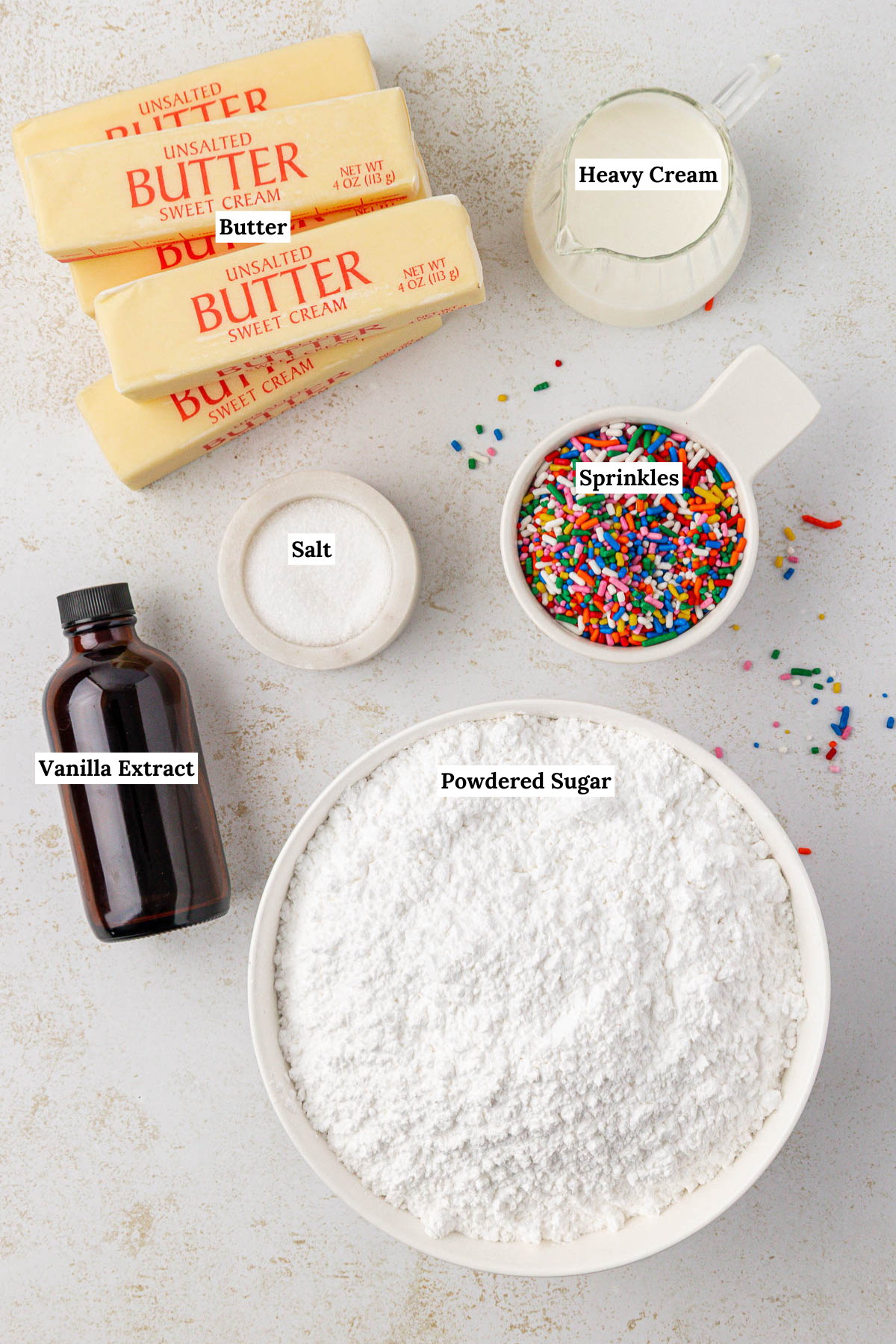 Top-down view of baking ingredients: four sticks of butter, a small pitcher of heavy cream, a bowl of sprinkles, a bottle of vanilla extract, a bowl of salt, and a large bowl of powdered sugar, all labeled.