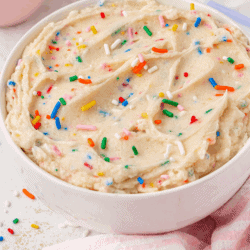 A bowl of fluffy, smooth frosting with colorful sprinkles sits next to a small dish of more sprinkles and a cake in the background. Text on the image reads, Fluffy & Smooth Funfetti Frosting.