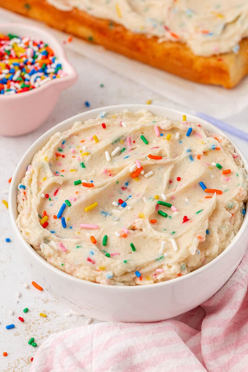 A white bowl filled with creamy funfetti frosting topped with colorful rainbow sprinkles sits on a white surface. A pink-striped cloth and a small bowl of extra sprinkles are next to it. A cake with matching frosting is in the background.