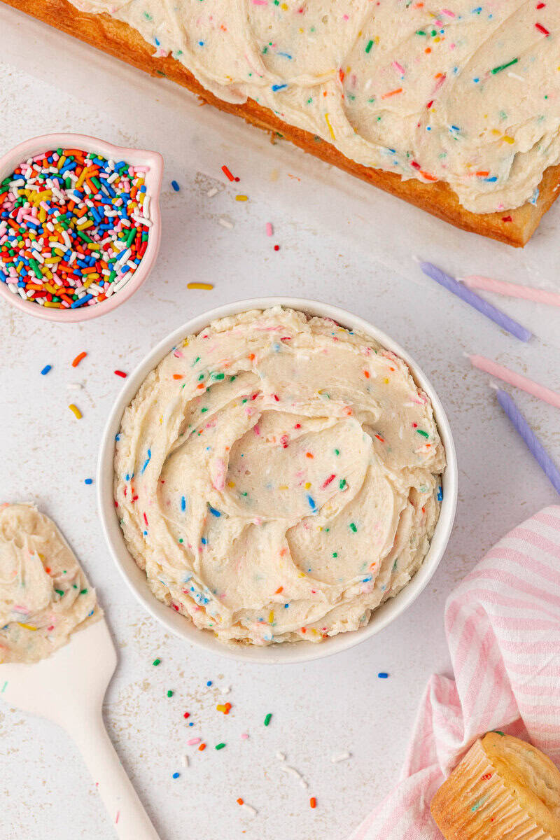 A bowl of funfetti frosting with colorful sprinkles sits on a white surface, surrounded by a spatula, a small bowl of extra sprinkles, pink and purple candles, and a partially frosted cake.