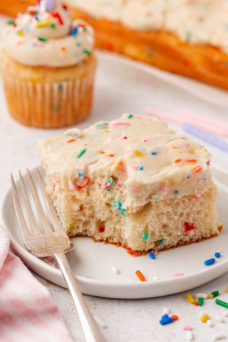A square slice of vanilla sheet cake with white frosting and colorful sprinkles sits on a plate with a fork. In the background, a cupcake and more cake are visible, both topped with similar frosting and sprinkles.