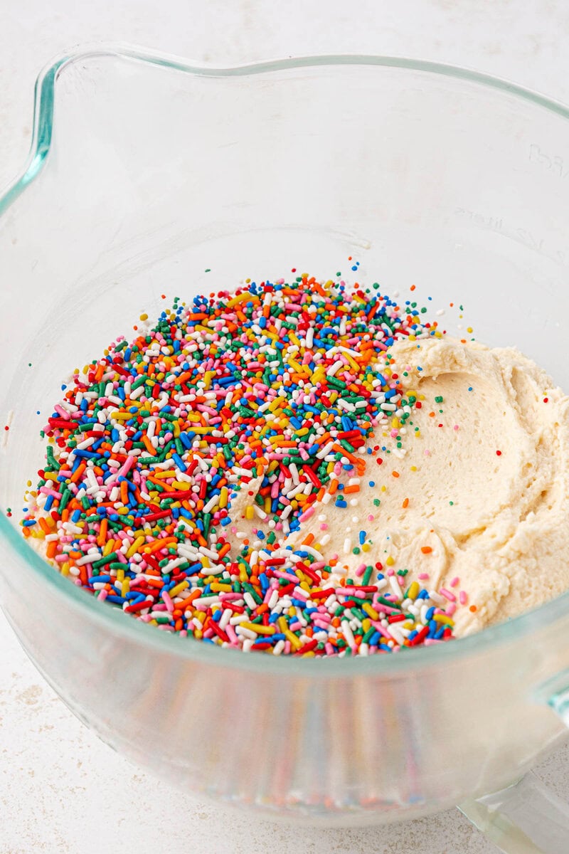 A glass mixing bowl with cookie dough and colorful rainbow sprinkles sitting on top, ready to be mixed together.