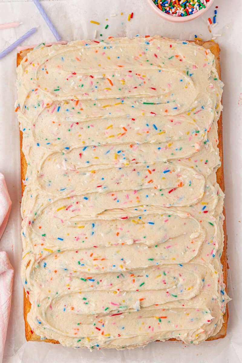 A rectangular sheet cake topped with swirled vanilla frosting and colorful rainbow sprinkles on parchment paper, with a bowl of extra sprinkles nearby.