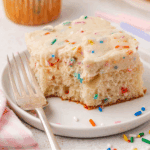 A slice of vanilla cake with colorful sprinkles and thick frosting sits on a white plate next to a fork. A few extra sprinkles are scattered on the plate.
