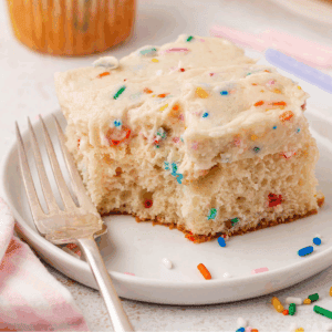 A slice of vanilla cake with colorful sprinkles and thick frosting sits on a white plate next to a fork. A few extra sprinkles are scattered on the plate.