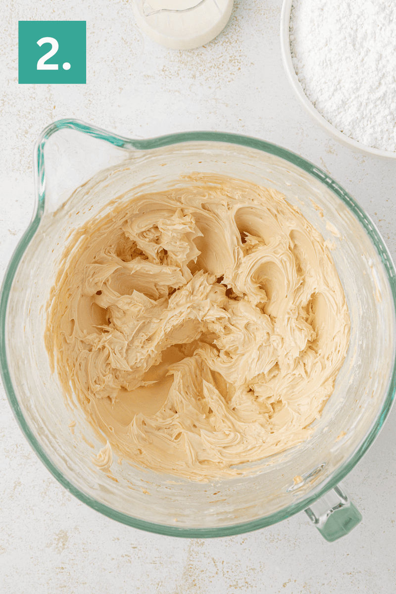 A glass mixing bowl filled with creamy, light brown butter mixture. A bowl of powdered sugar and a small bottle of liquid are nearby. A green square in the corner displays the number 2.