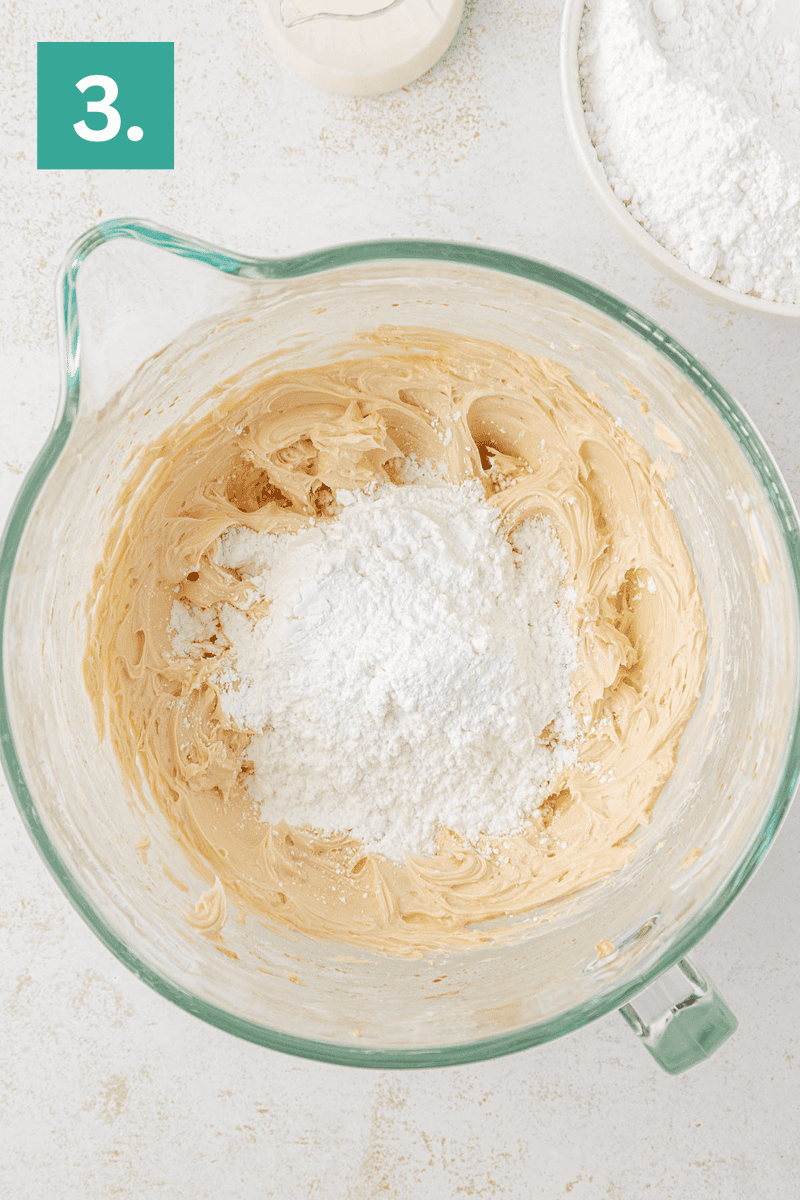 A glass mixing bowl with creamy batter and a heap of powdered sugar in the center. Part of a white bowl with flour and a bottle of milk are nearby. A green box with the number 3 appears in the upper left corner.