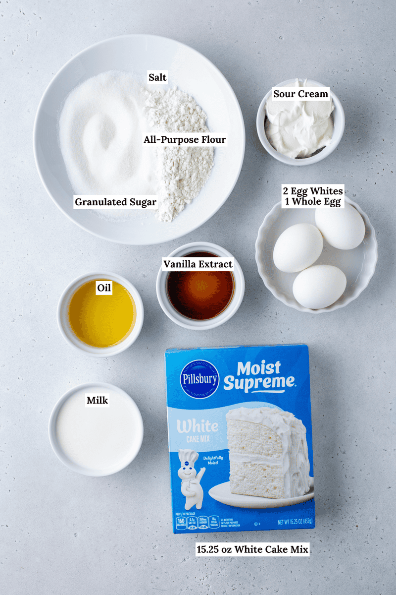 Top-down view of cake ingredients in bowls on a light surface, including flour, sugar, salt, sour cream, eggs, oil, milk, vanilla extract, and a box of Pillsbury Moist Supreme White Cake Mix, all labeled.