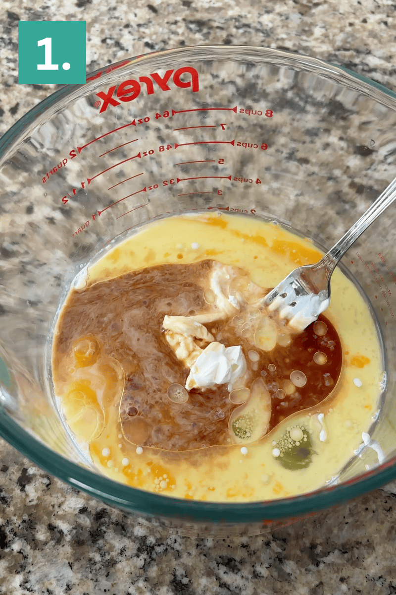 A glass Pyrex measuring cup on a granite countertop contains eggs, vanilla, melted butter, and sour cream being mixed with a fork. A green box in the corner displays the number 1.
