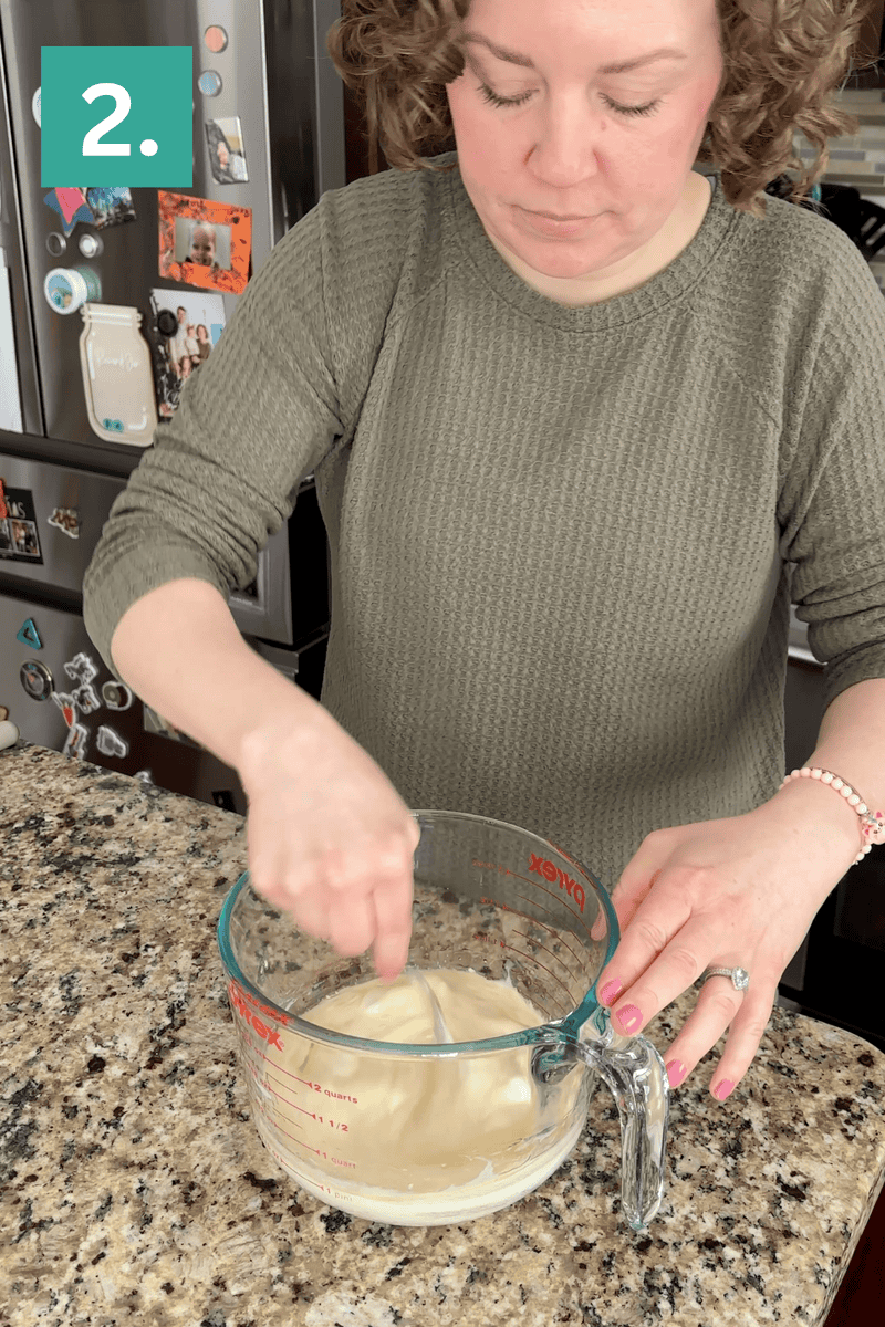 A person in a green sweater stirs dough in a large glass measuring cup on a granite kitchen counter. The number 2. is shown in the top left corner of the image.
